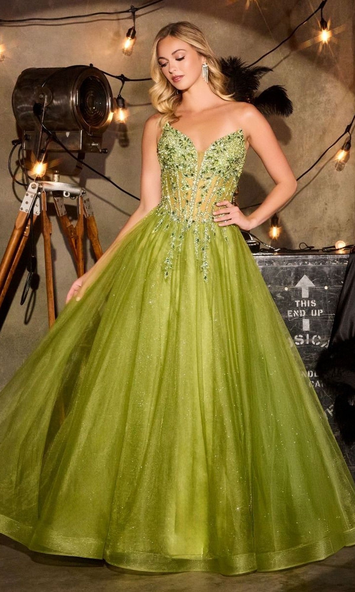 A woman in the Lace-Up Strapless Prom Ball Gown: Ladivine CD721—featuring sparkling green glitter tulle, a beaded corset bodice, and a full skirt—stands indoors among string lights and vintage decor.